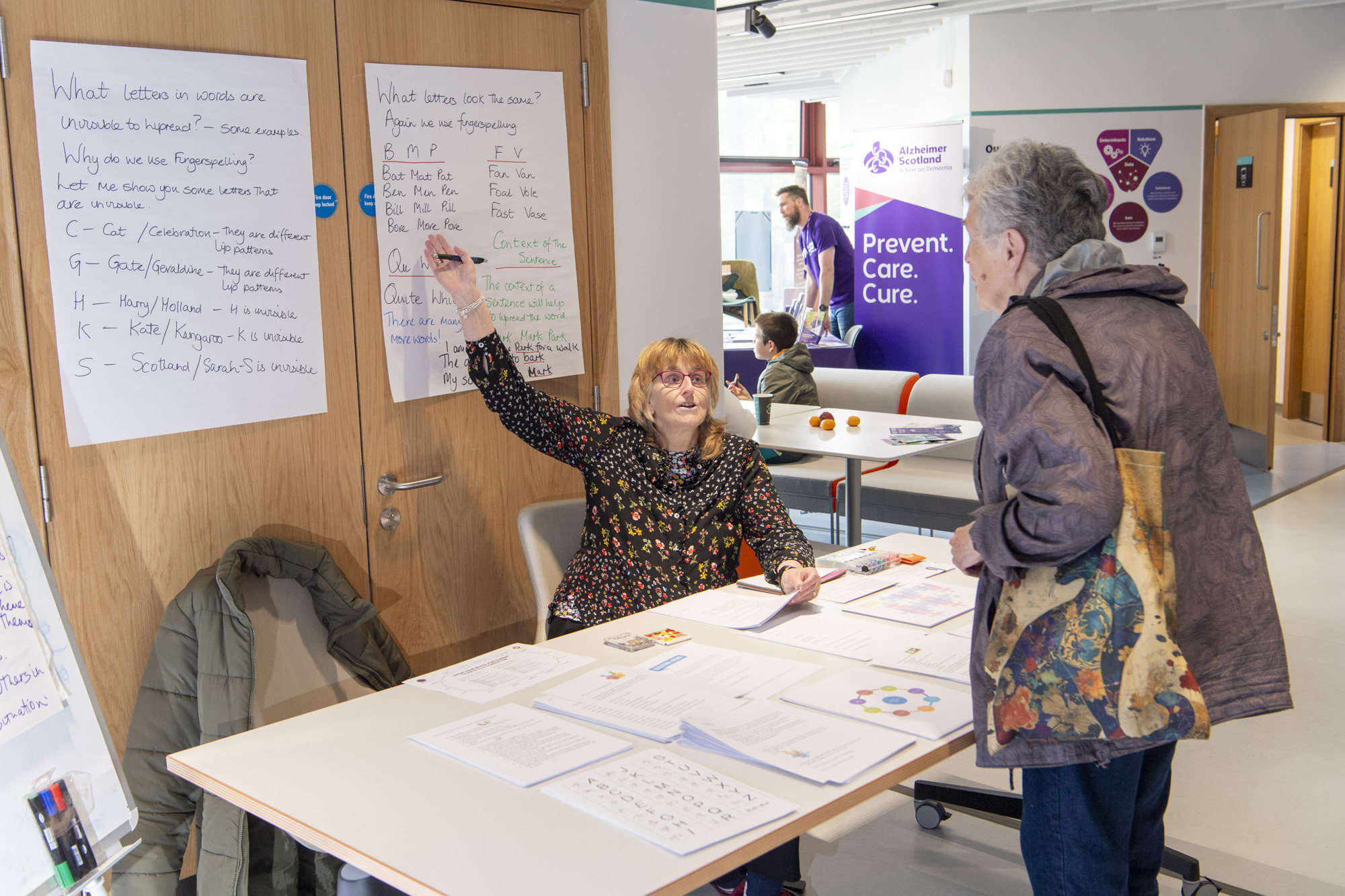 Woman sitting at information table with educational materials and posters, another woman is standing looking at the materials