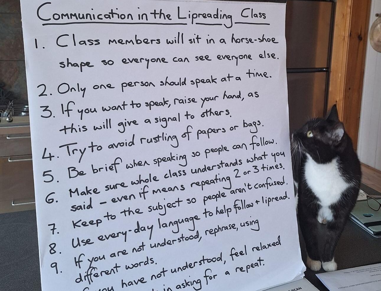 Handwritten list of communication guidelines for lipreading class with black and white cat sitting beside it