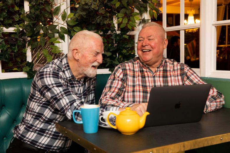 Two older men laughing together while looking at a laptop, sitting at a table with coffee mugs and a yellow teapot