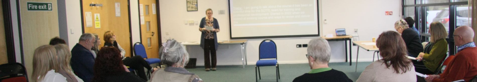 A group of people sit in a semicircle in a conference room, facing a speaker who stands next to a projected screen.