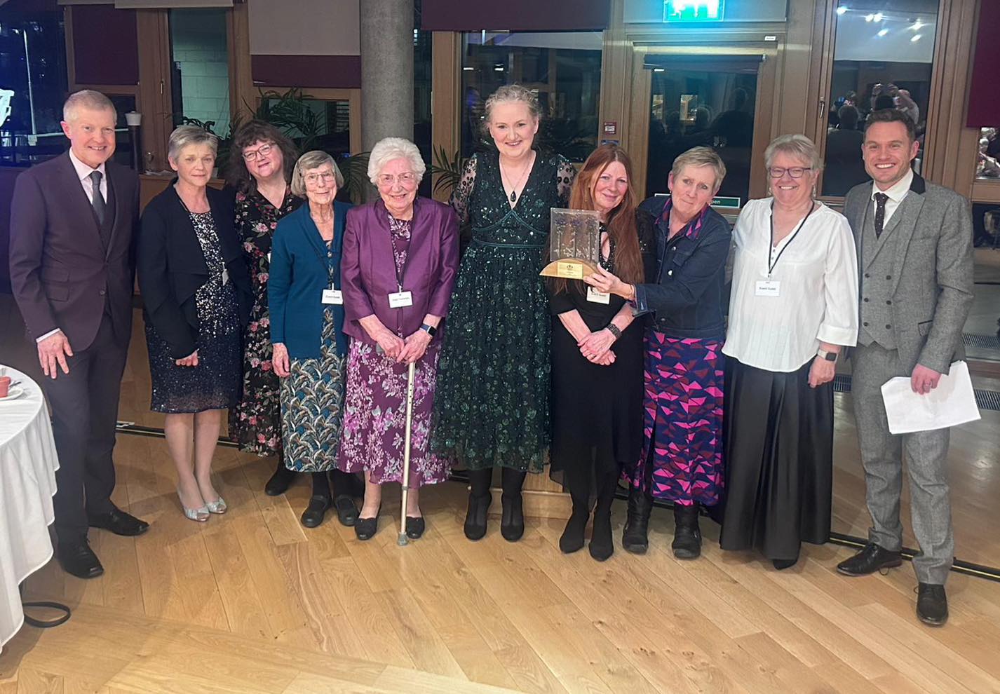 A group of nine people dressed formally pose indoors at an evening event. A person in a dark green sparkly dress stands in the center while another person beside them holds a framed award or plaque. The room has wooden floors, large windows, and warm lighting.